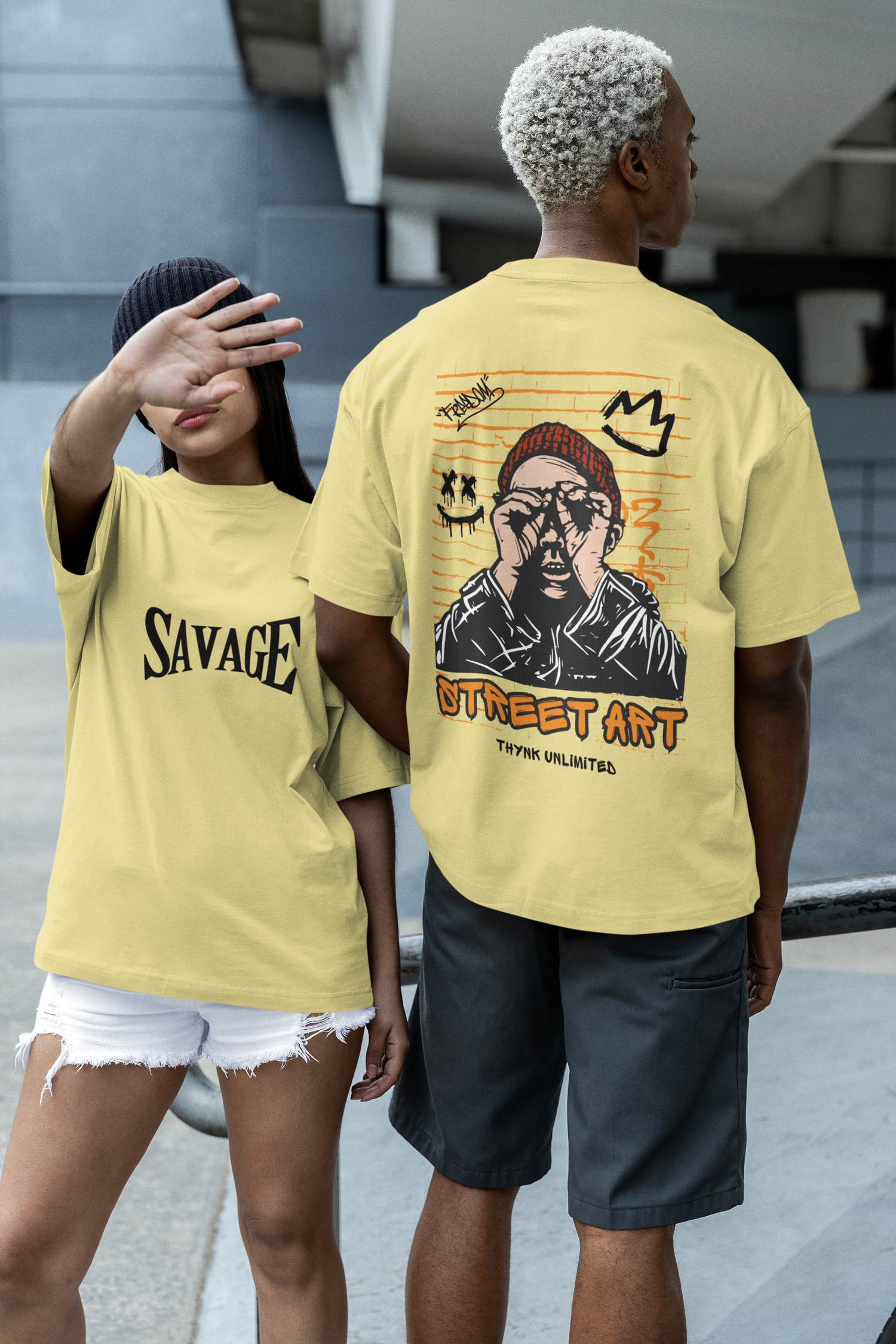 A girl wearing a beige oversized T-shirt featuring bold 'Savage' typography on the front, paired with a boy in a matching beige oversized T-shirt showcasing the 'Street Art' graphic design on the back, creating a stylish and coordinated streetwear look by Looma Fashion.
